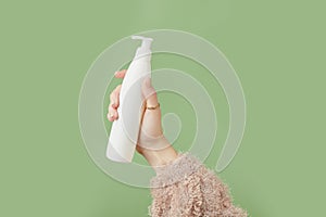A womans hand in a sweater holds a bottle of cream on a green background. Self care beauty treatment