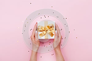 Womans hands holding gift or present box decorated confetti on pink pastel table top view. Flat lay composition for birthday.