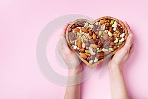 Womans hands holding heart shaped bowl with mixed nuts on pink table top view. Healthy food and snack. Flat lay