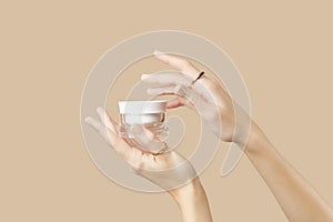 A womans hands holding a white cosmetic jar on a beige background. Beauty treatment concept