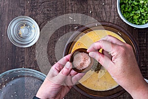 Womanâ€™s hands adding a pinch of pink Himalayan salt to raw egg mixture in glass bowl, on a wood table