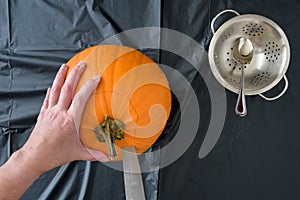 Womanâ€™s hands caving fresh pumpkin on a plastic covered table, knife, spoon, and bowl