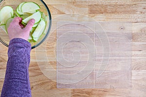 Womanâ€™s hands taking granny smith apple slices out of a glass bowl and laying them out on a mesh tray for dehydrating