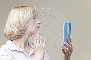 Women Ageing Concepts. Mature Caucasian Woman Examining Her Face In Hand Mirror And Examining Skin Indoors