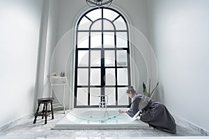 Women in bathrobe while carryin bathbomb in nice design jacuzzi bathtub with high transparant window in natural light setting