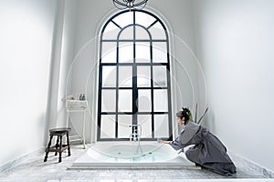 Women in bathrobe while carryin bathbomb in nice design jacuzzi bathtub with high transparant window in natural light setting photo