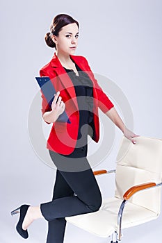 Women in Business Ideas. Portrait of Caucasian Brunette Enterpreneur in Red Blazer Posing At Chair With Paper Folder Notebook
