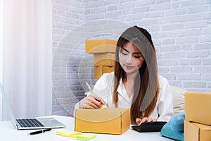 Women business owner writing address on packing box at workplace in home offce. online shopping SME entrepreneur or freelance