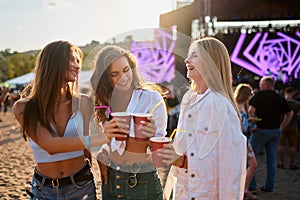 Women clink cups at beach music fest, happy friends toast with drinks in sunshine. Outdoor party vibe, casual summer fun