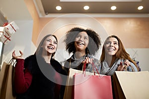 Women of diverse ethnicity with shopping bags posing in mall on sale. Portrait of three smiling multiracial girls look