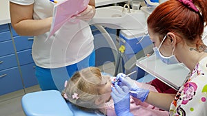 Women doctor in medical mask using tools for cures teeth of little patient and beside assistant