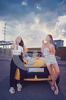 Women enjoying soda in glass bottles and eating french fries, leaning on trunk of yellow car with pizza on it. Fast food