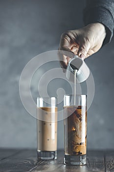 Women hand is pouring homemade sour cream from small jar to glass with brew cold coffee and ice. Cold summer drink on a dark