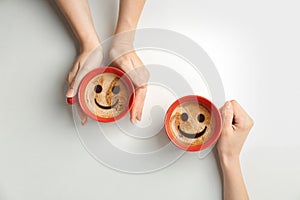 Women holding cups of delicious hot coffee with foam and smiles on light background, top view.