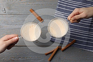 Women holding glass cups of delicious eggnog with cinnamon at grey wooden table, top view
