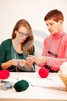 Women knitting with red wool. Eldery woman transfering her knowledge of knitting on a younger woman on handcraft workshop.