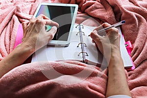 Women laying in bad covered by pink plaid. With tablet, pen and pink notebook. Working at home. Quarantine