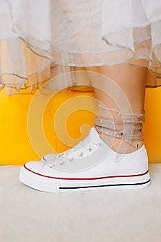 Women legs in white clean new sneakers, transparent thin socks with silver shiny stars and lush tulle dress on yellow background.