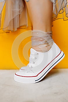 Women legs in white clean new sneakers, transparent thin socks with silver shiny stars and lush tulle dress on yellow background.