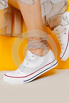 Women legs in white clean new sneakers, transparent thin socks with silver shiny stars and lush tulle dress on yellow background.