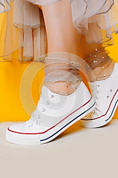 Women legs in white clean new sneakers, transparent thin socks with silver shiny stars and lush tulle dress on yellow background.