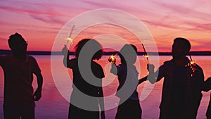 Women and men with sparklers having fun on lake beach dancing in darkness