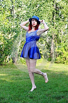 Women with red curly hair in a classic blue short dress and blue hat, jumping on one leg on the green lawn