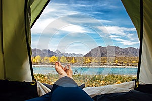 Women`s bare feet in blue trousers crossed on the edge of green tourist tent. Turquoise river against rocky mountains and blue sk