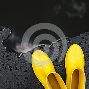 Women`s bright yellow rubber boots stand under raindrops on a black background in front of a puddle.