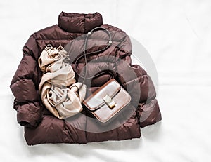 Women`s brown down jacket, cashmere scarf, leather shoulder bag on a light background, top view. Autumn, winter women`s clothing