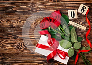 Women`s day background. 8 march wooden calendar, red roses, gift with red ribbon on the wooden background. Copy space, flat lay