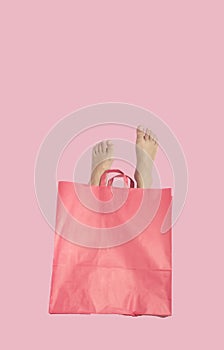 A women`s feet come out of a paper pink shopping bag on a pastel pink background. Surreal shopping background.