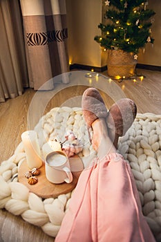 Women`s feet in home fur slippers, cup of cappuccino, candles on blanket of thick yarn and Christmas tree with lights