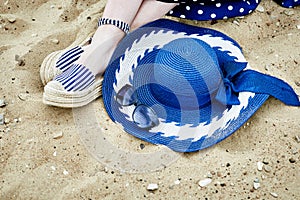 Women`s feet in striped sandals, blue hat and sunglasses on the sand