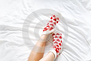 women& x27;s feet in warm socks with red hearts on a white bed