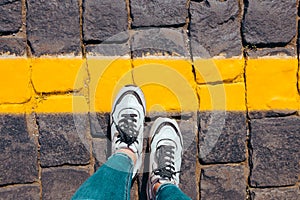 Women`s feet in white sneakers stand on the pavement one foot stepped on a bright yellow line, cross the line