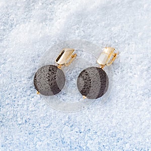 Women`s golden earrings with round black volcanic lava stone on white snow background