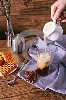 Women`s hand with creamer pouring milk in glass with coffee. Cinnamon sticks, homemade cookies and bunch of wildflowers