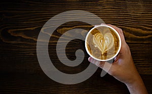 Women`s Hand holding cup of cappuccino coffee with heart shape latte art on old wood table Background, Latte coffee on old wooden