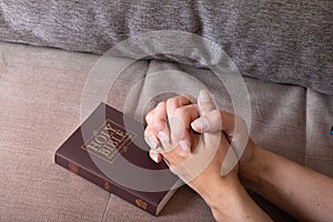 Women's hands folded in prayer over the Holy Bible at home sofa