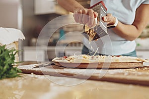 Women& x27;s hands grate cheese on a pizza lying on the table in the background of the kitchen