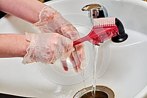 Women& x27;s hands in hair coloring gloves wash a comb under a stream of water in a white sink