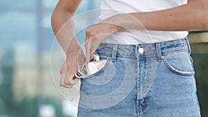 Women`s hands hide smartphone tangled white headphones in jeans pocket outdoor. Girl tries untangle earbuds, roll wires