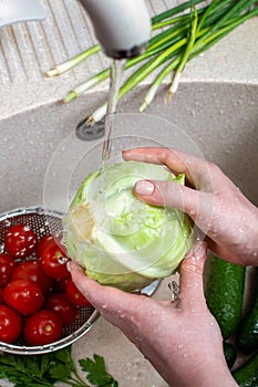 Women`s hands hold a fresh head of cabbage under a stream of water from the tap over the kitchen sink where there are fresh