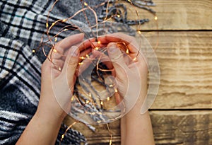 Women `s hands hold a garland of Christmas lights .Warm grey scarf or plaid on a wooden background