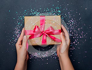 Women`s hands hold a gift box with a pink satin bow on a dark gray background. Sparks are scattered around