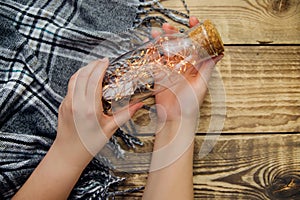 Women `s hands hold a glass bottle with a garland of Christmas lights .Warm grey scarf or plaid on a wooden background