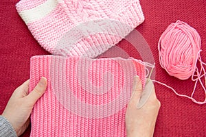 Women`s hands hold a knitted pink cloth, which is tied with a crochet. Nearby is a children`s hat tied with spokes