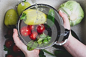 Women`s hands hold a metal colander full of fresh ripe vegetables and herbs under a spray of water over the kitchen sink where th