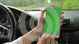 Women`s hands hold a phone with a green screen with their hands on the steering wheel of the car. The girl driver is sitting at th